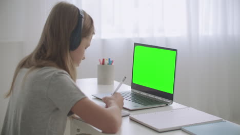 e-learning-for-schoolers-by-internet-girl-is-drawing-in-copybook-listening-teacher-by-videochat-green-screen-on-laptop
